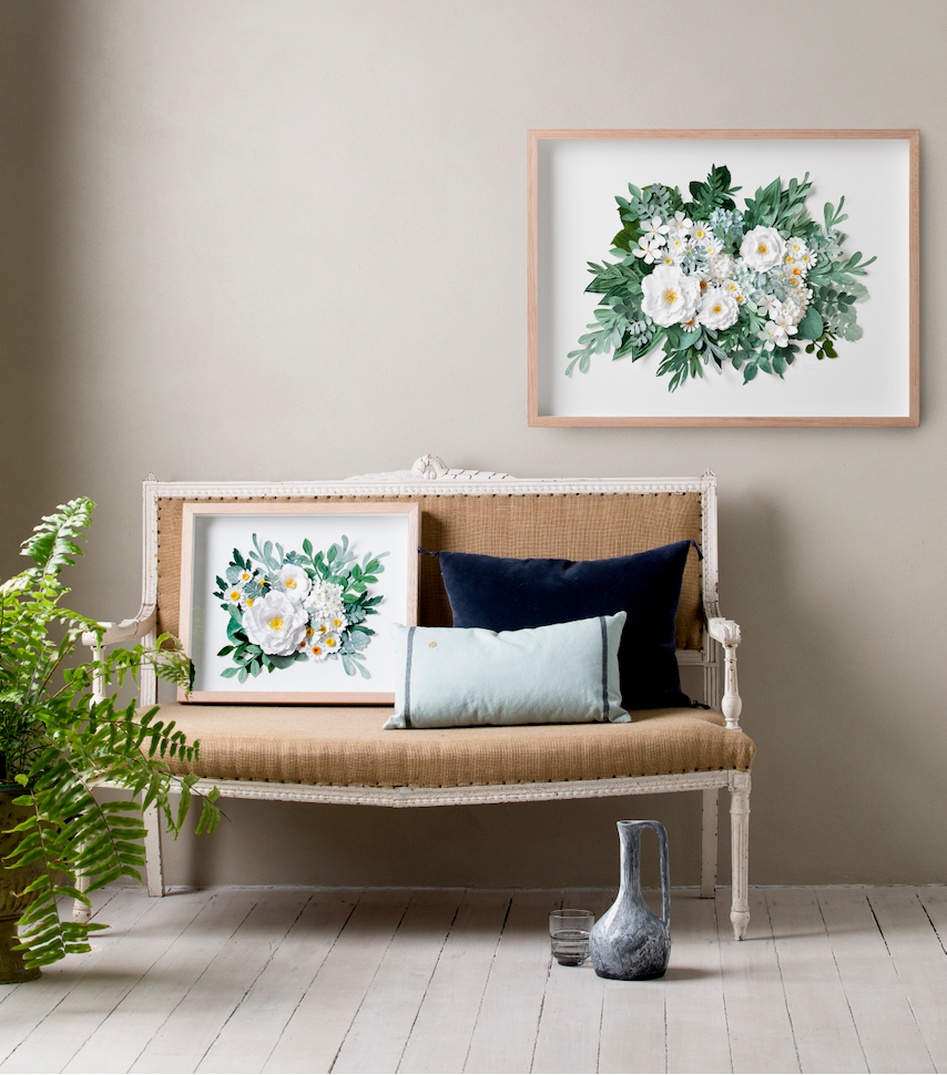 A styled interior scene with a beige upholstered bench against a warm taupe wall, featuring two framed floral watercolor prints. One print is hung on the wall, and the other rests on the bench.