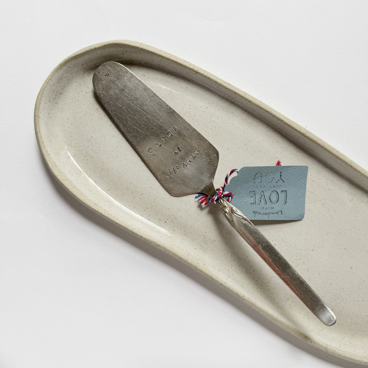 A rustic metal cake/pie server with engraved text, resting on a matching speckled ceramic serving tray. The server is tied with a small blue paper tag that reads 'LOVE' and has a red-and-white string accent.