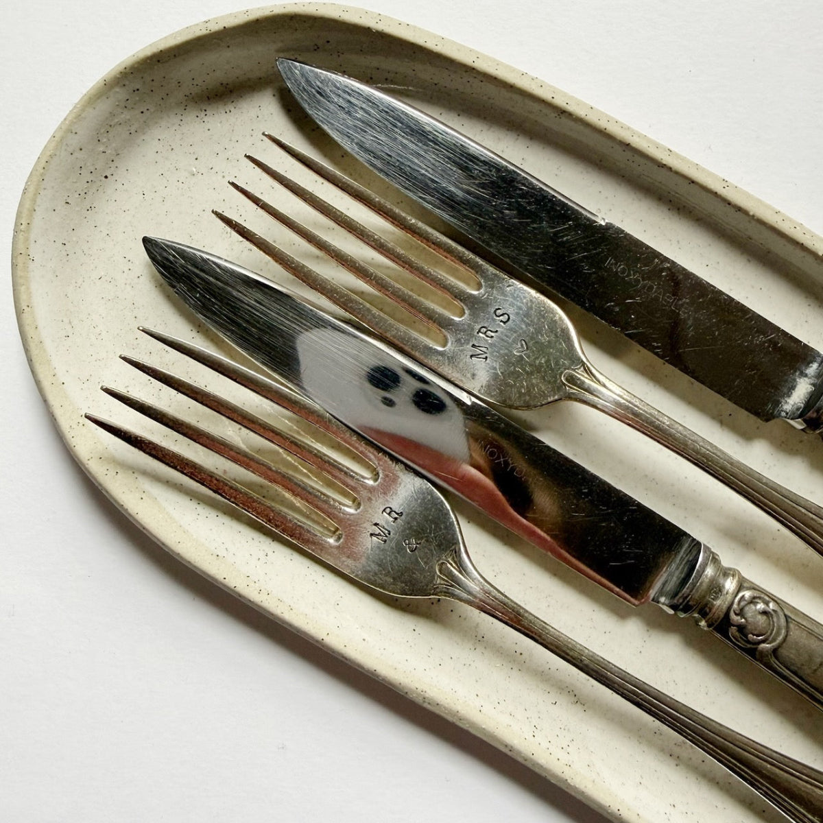 A close-up of three engraved silver forks and knives arranged diagonally on a speckled beige ceramic oval plate against a clean white background.