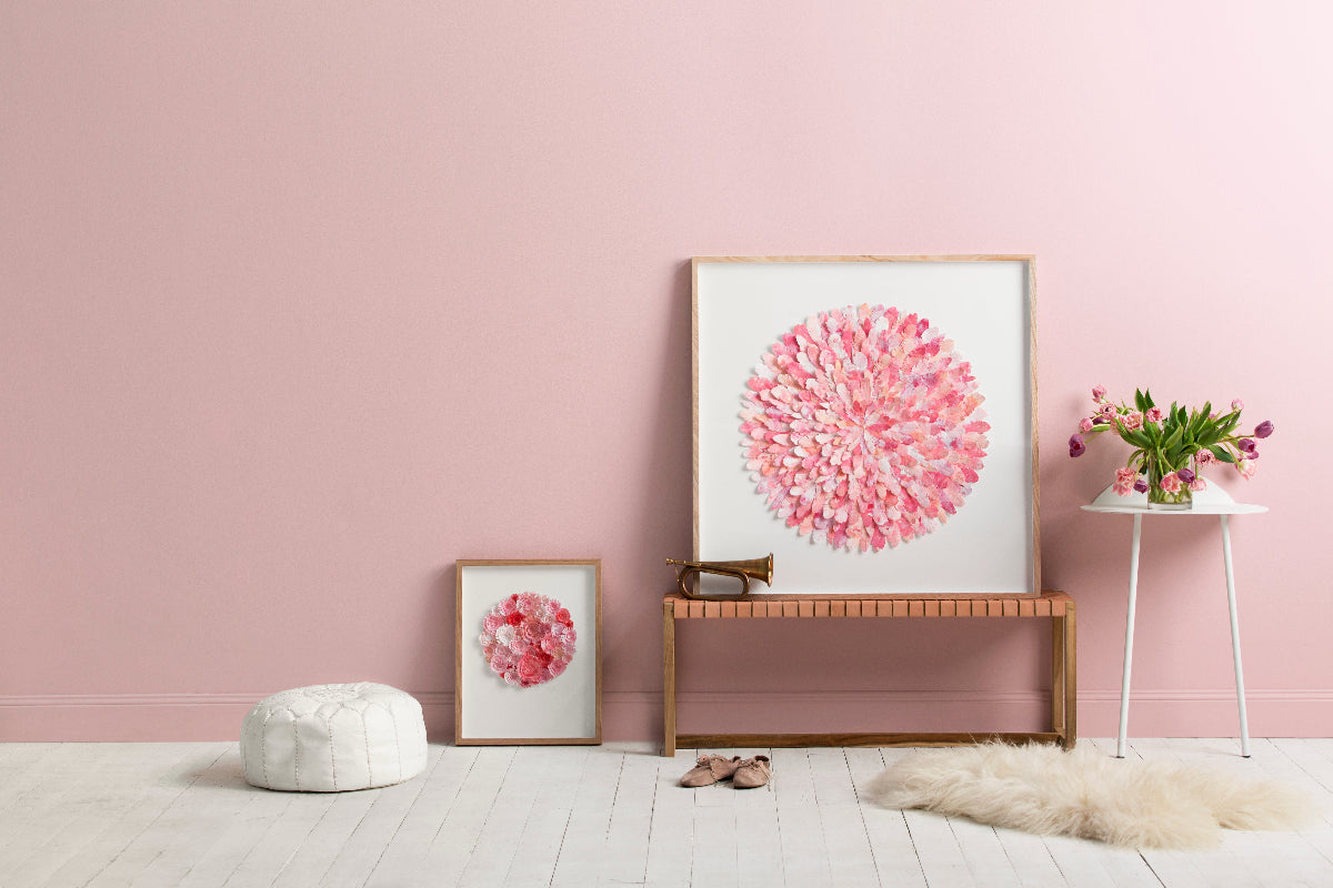 A styled interior scene with a large framed pink floral artwork leaning on a wooden bench, a smaller matching framed piece on the floor, a white pouf, a white side table with flowers, and a cream rug against a pink wall.