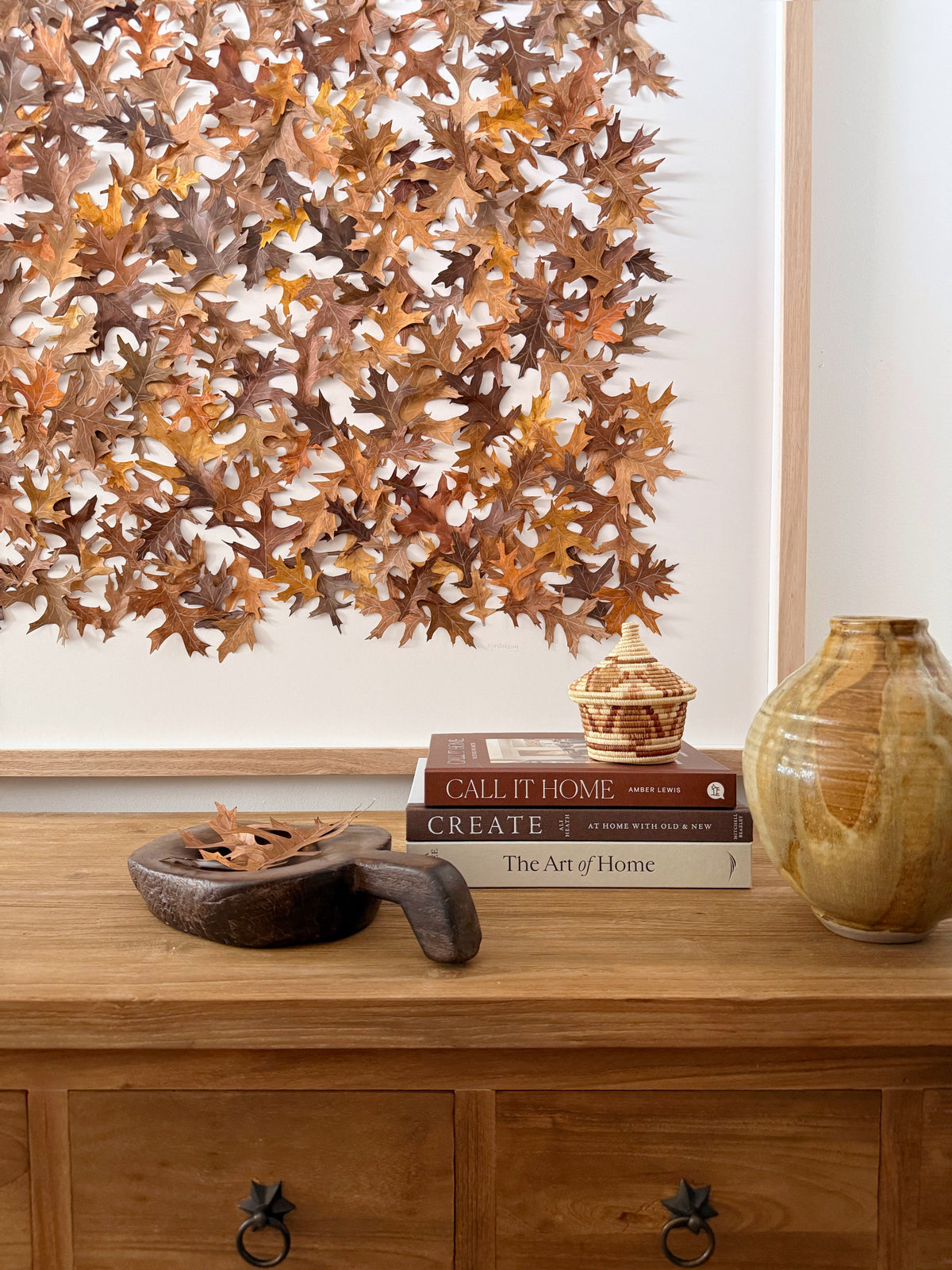 Decorative wall art with autumn leaves above a wooden console table with books and a vase.