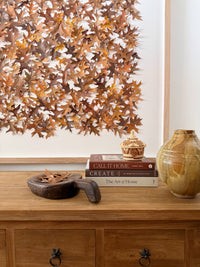 Decorative wall art with autumn leaves above a wooden console table with books and a vase.