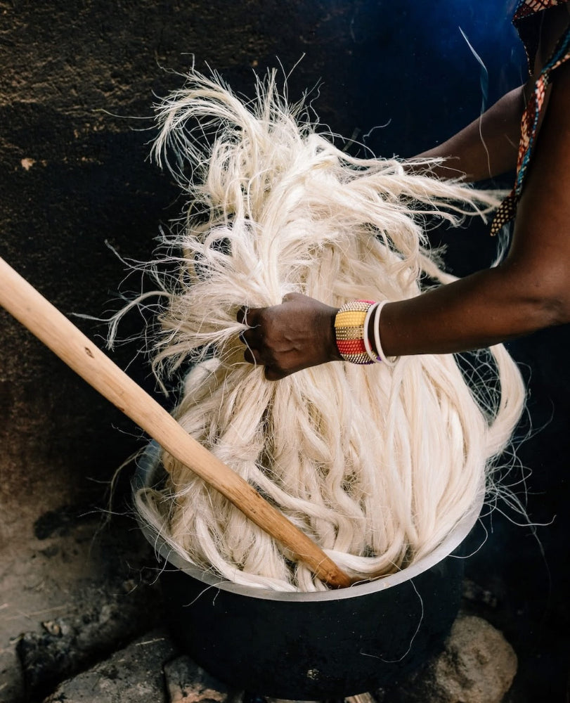 A person's hand and arm, adorned with colorful bracelets, holds a large bundle of off-white raw fiber or yarn inside a dark metal pot, with a wooden stirring rod resting inside.