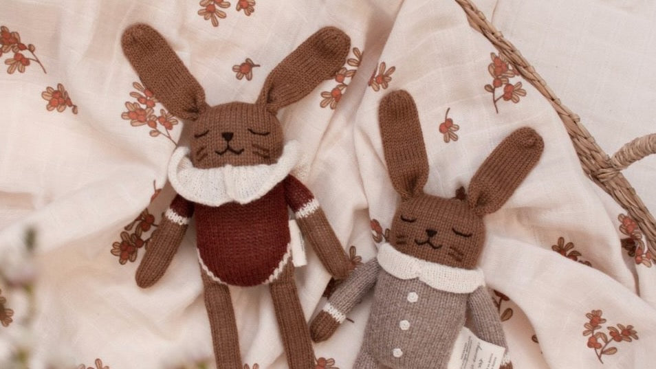 Two knitted rabbit toys lying on a cream fabric backdrop with a floral print. One rabbit is brown with a white ruffled collar and a dark brown sweater.
