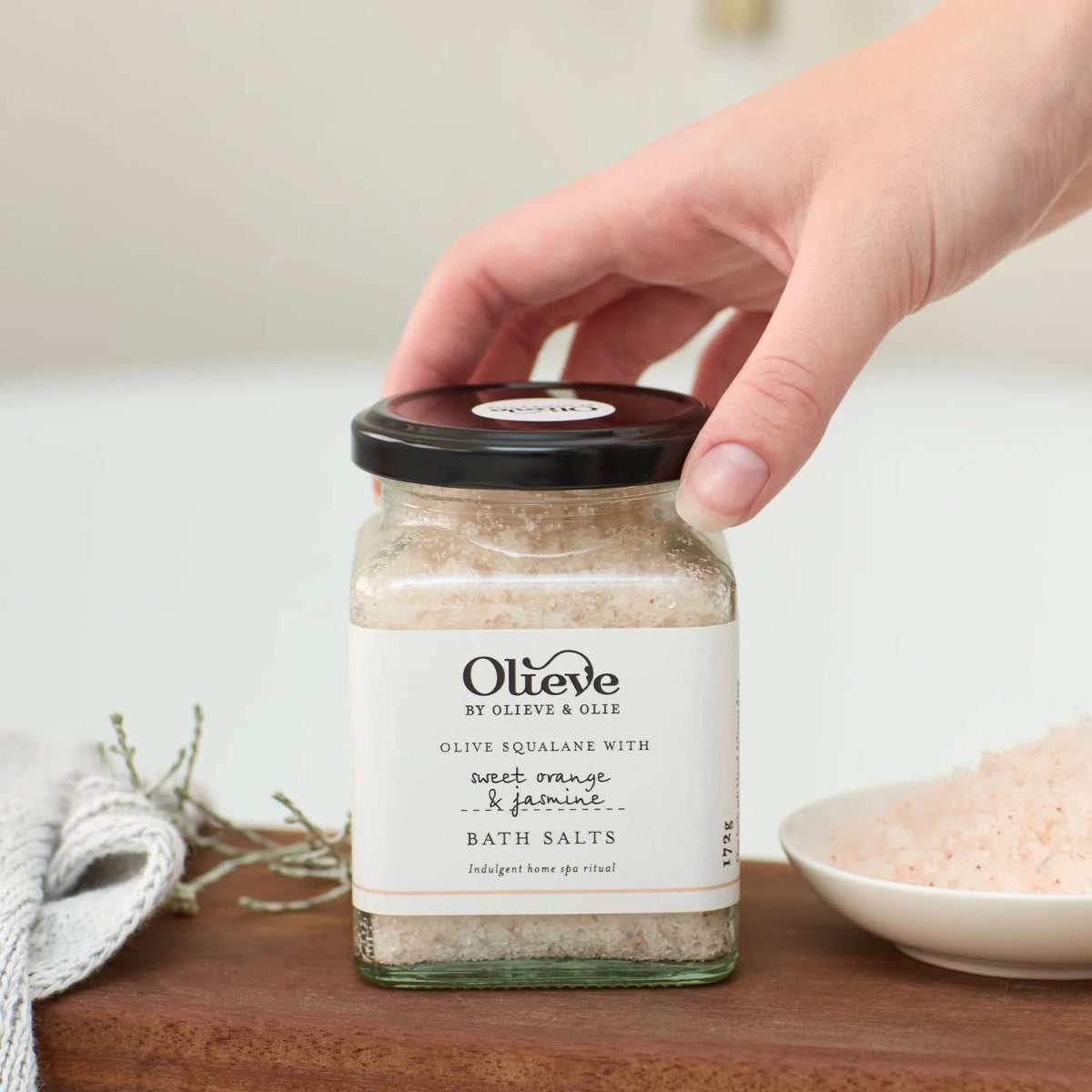 A hand holding the lid of a glass jar of Olieve Bath Salts on a wooden tray, with a bowl of pink bath salts and a soft bathroom background.