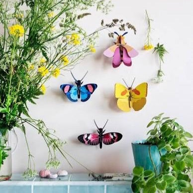 Colourful paper butterflies on a white wall near plants.